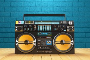 A retro black boombox with yellow speakers sits on a wooden floor against a vibrant blue brick wall. The boombox, complete with various buttons, dials, and displays, stands as resilient as someone job hunting in today's market. A rectangular handle is fixed on top, reminiscent of 1980s portable stereo systems.