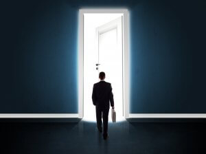 A person holding a briefcase and wearing a suit walks towards a bright, open door in a dark room. Light pours through the doorway, illuminating the figure and casting long shadows on the floor. The scene evokes a sense of opportunity, transition, and being promoted to new possibilities.