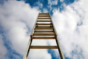 A wooden ladder, promoted by its low-angle perspective, extends upwards toward a sky filled with white, fluffy clouds, giving the impression that it reaches into the heavens.