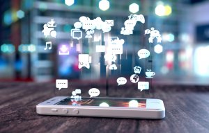 A smartphone rests on a wooden surface with numerous social media and app icons floating above it, including chat bubbles, music notes, shopping carts, and game controls, depicting a vibrant digital interaction. The background is blurred, suggesting a cityscape at night.