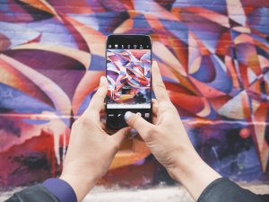 A person holding a smartphone capturing a photo of a colorful graffiti mural on a brick wall. The mural features abstract designs with swirling patterns in red, purple, blue, and orange hues. The person's fingers are visible gripping the phone.