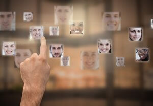 A hand with the index finger extended is touching a digital interface displaying floating profile pictures of diverse people. The background is blurred, placing focus on the photos and the hand interacting with them.