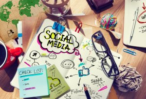 A cluttered workspace features a notebook with a SOCIAL MEDIA brainstorm, sticky notes, a map, a red coffee mug, eyeglasses, and various office supplies. Drawings and keywords like "Share" and "Tweet" visualize social media strategies.