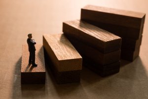 A miniature figure of a man in a suit stands on a wooden block, looking up at a staircase made of wooden blocks that ascend in size from left to right. Under warm, dramatic lighting, the scene emphasizes career growth and personal development as he eyes the next vacancy awaiting him.