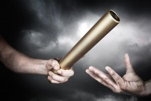 Close-up of two hands in front of a dark, cloudy sky. The left hand is passing a gold baton to the right hand, symbolizing the act of passing control or responsibility, filling a vacancy with leadership and purpose.