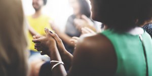A group of people in casual attire are clapping and smiling, creating a lighthearted and supportive atmosphere. The image focuses on one team member's hands in the foreground, clapping, with others visible in the background slightly out of focus.