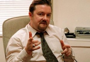 A man with a mustache and beard, wearing a light-colored shirt and patterned tie, sits in an office chair. He is gesturing with both hands, pointing his fingers outward. Behind him are an office window with blinds and some desk items typical of office bosses.