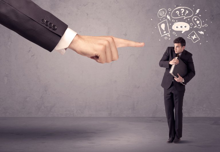 A large hand points at a nervous man in a suit holding documents as he stands against a grey wall. The man appears confused, with various question marks and symbols drawn above his head, suggesting uncertainty or stress—possibly from the demands of bosses.