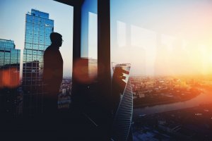 Silhouette of a boss standing by a large window in a high-rise building, overlooking a cityscape at sunset. The glass reflects nearby skyscrapers and soft, warm lighting enhances the tranquil ambiance.
