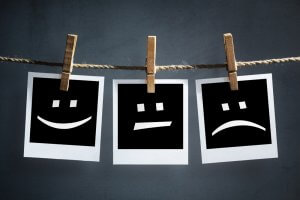 Three black-and-white squares with different facial expressions (smiling, neutral, and frowning) are hanging on a clothesline with wooden clothespins against a gray background—an artistic depiction of customer service experiences.