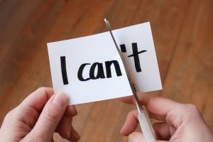 A person is using a pair of scissors to cut through a piece of paper that has the words "I can't" written on it, effectively cutting off the "n't" to change the message to "I can," symbolizing their determination to overcome fear. The background is a wooden surface.