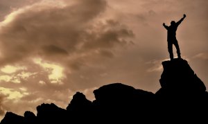 Silhouette of a person, who has overcome fear, standing triumphantly with arms raised on the peak of a rocky mountain, against a dramatic, cloudy sky at sunset.