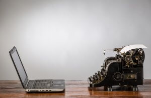 A modern laptop and a vintage typewriter sit side by side on a wooden table against a gray background, illustrating the contrast between old and new technology. The typewriter has a sheet of paper inserted, while the laptop, angled to the left, epitomizes modern information governance.