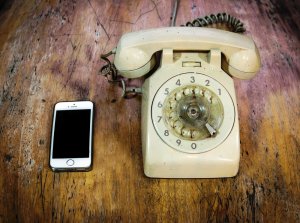 An old, beige rotary dial telephone with a curly cord is placed next to a modern smartphone on a rustic wooden surface, symbolizing the evolution of information governance. The smartphone has a blank, black screen.