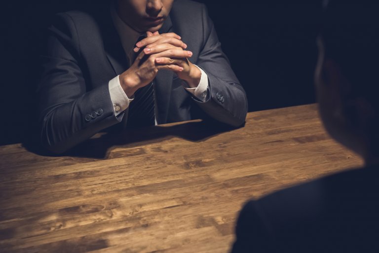 A person in a suit sits at a wooden table with their hands clasped under their chin in a dimly lit room, looking intently across the table. Another individual, partially visible in the foreground, is seated opposite. The scene suggests an intense interview.