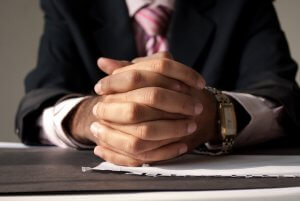 Close-up of a person wearing a suit and striped tie with their hands clasped together on a table, ready for an interview. A stack of papers and a partially visible watch on the person's wrist are also in view. The background is out of focus.