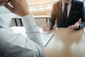 Two individuals sit across from each other at a table, possibly in an interview setting. One person is holding a clipboard with documents, while the other, dressed in a suit and tie, gestures with their hands as they discuss.