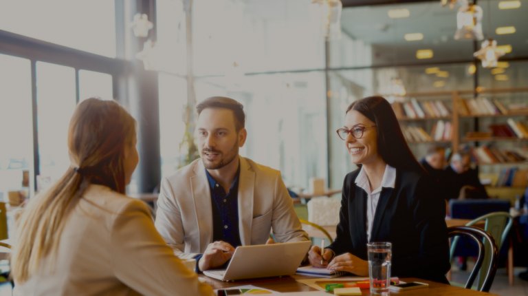 Three people sit around a table, engaged in a business discussion. Two are sitting opposite one individual, with a laptop open in front of them. Dressed in professional attire, they appear to be having a productive conversation about the interview process in a well-lit office space.