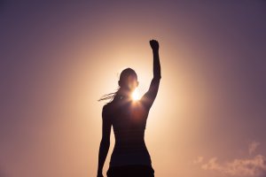 Silhouetted against a glowing sunset, a person stands with their arm raised triumphantly in the air. The dramatic lighting highlights the figure's strong, confident pose, embodying self-belief and victory. The background is a gradient of warm hues from the setting sun.