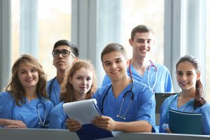 A group of six young medical professionals from Stanislaus County, wearing blue scrubs and stethoscopes, stand together indoors. Some hold clipboards and notebooks. They are smiling and looking at the camera, suggesting a positive and collaborative environment.