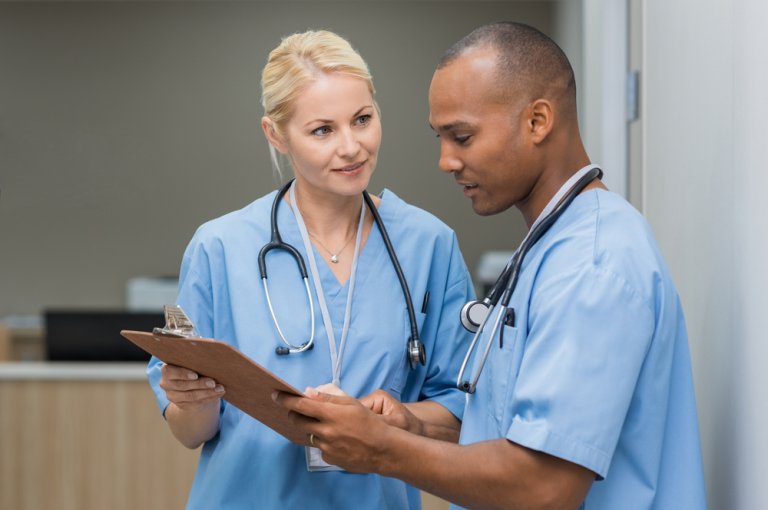 Two healthcare professionals in blue scrubs are discussing a document on a clipboard. The woman on the left is smiling and listening, and the man on the right is attentively explaining something, holding the clipboard. Both have stethoscopes around their necks, working diligently to serve Stanislaus County.