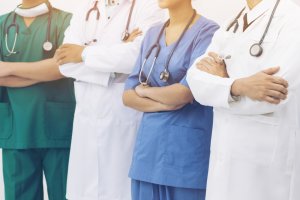 A group of four healthcare professionals stands with arms crossed in Stanislaus County, each wearing scrubs or lab coats with stethoscopes around their necks. The attire includes green, blue, and white variations, indicating different medical roles or departments.