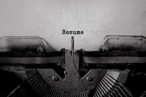 A close-up of an old-fashioned typewriter typing the word "Resume" on a piece of white paper. The typewriter keys and the roller holding the paper are visible, giving the image a vintage feel. The centered word "Resume" appears in classic, black, typewriter-style font.