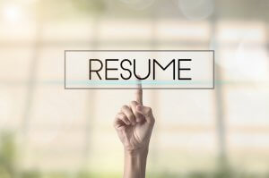 A hand pointing upwards towards a glass surface with the word "RESUME" displayed on it. The softly blurred background suggests a modern, light-filled environment, conveying themes of resume-building and career development.