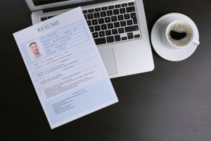 A resume on a light blue sheet of paper lies on an open silver laptop keyboard. To the right of the laptop sits a cup of black coffee on a dark wooden table. The resume, detailing impressive credentials, includes a small, square photo of the applicant in the top left corner.