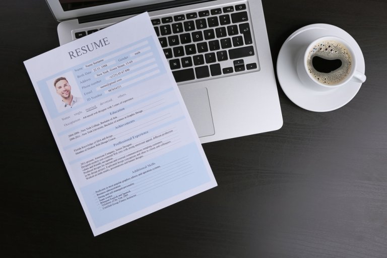 A resume on a light blue sheet of paper lies on an open silver laptop keyboard. To the right of the laptop sits a cup of black coffee on a dark wooden table. The resume, detailing impressive credentials, includes a small, square photo of the applicant in the top left corner.