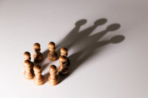 Six wooden chess pawns arranged in a circular formation on a white surface. The pawns cast shadows that converge and overlap, creating the illusion of two taller chess figures in the shadows. With self-belief, these small pieces depict how unity and perspective can elevate even the humblest elements.