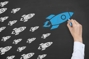 A person's hand, holding blue chalk, draws a blue rocket ship on a blackboard. Several smaller white rocket ships, drawn in chalk, are positioned behind it, suggesting the blue rocket is leading them. The scene symbolizes leadership and self-belief in driving innovation.