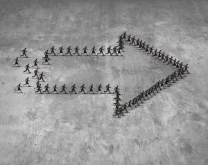 A group of people in business attire standing on a concrete surface form the shape of a large, right-pointing arrow. Smaller groups of people, reflecting shared values, create the left side and tail of the arrow, converging towards the tip. The ground is textured and gray.