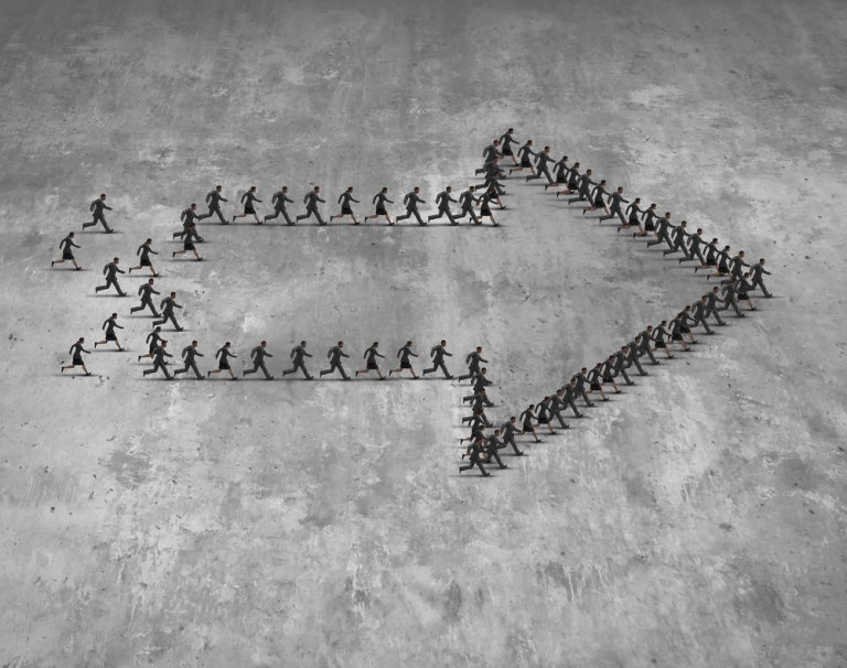 A group of people in business attire standing on a concrete surface form the shape of a large, right-pointing arrow. Smaller groups of people, reflecting shared values, create the left side and tail of the arrow, converging towards the tip. The ground is textured and gray.