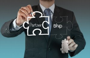 A businessman in a suit is holding two puzzle pieces. The left piece, labeled "Partner," is being connected with the right piece, labeled "ship," completing the word "Partnership." This signifies forming a business alliance or collaboration grounded in shared values.