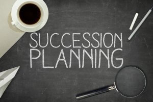 A chalkboard with "Succession Planning" carefully written in white chalk. The board also features a white paper airplane, a coffee cup, a piece of white chalk, a pen, and a magnifying glass.