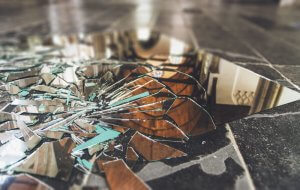 Broken mirror shards scattered on a dark, polished floor. The reflective pieces create a fractured and distorted reflection of the interior space, including blurred images of furniture and architectural details. Evoking both disarray and fragmentation, the scene whispers age-old superstitions about shattered luck.
