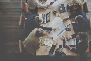A team of people in business attire sit around a wooden table, engaged in a meeting. Documents, laptops, tablets, coffee cups, and snacks are on the table. One person is pointing at a printed document, and others are looking at laptop screens or holding papers.