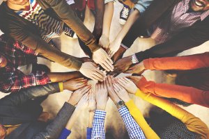 A diverse team of people stand in a circle with their arms extended, placing their hands on top of each other in the center. The image captures the unity and teamwork among the group, with various clothing styles and colors visible.