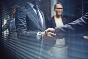 Two businesspeople shaking hands in a professional setting, with blurred figures of other employees in the background. They are dressed in formal business attire, suggesting a corporate environment or a business meeting.