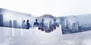 A handshake superimposed over a city skyline with several silhouettes of employees interacting and conversing within the arms. The image symbolizes teamwork, collaboration, and successful business partnerships amidst an urban backdrop.