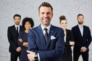 A smiling man in a blue suit stands confidently with his arms crossed in the foreground. Four business professionals, two women and two men, stand behind him, all dressed in formal attire against a white brick wall. The group of employees appears positive and united.
