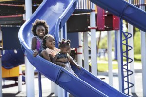 Three children are enjoying a slide at a playground. The first child is smiling widely at the top, while the other two are sitting close together on the slide, giggling as they descend. The playground equipment is colorful, and it is a sunny day.