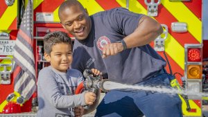 A firefighter joyfully assists a young boy in operating a fire hose, spraying water. They are standing in front of a brightly colored fire truck with red and yellow stripes. The boy looks excited as he holds the hose, while the firefighter enthusiastically guides him.