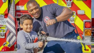 A firefighter helps a smiling young boy spray water from a fire hose in front of a fire truck with bright yellow and red stripes.