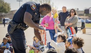 A police officer leans down to help a group of children seated on the ground while holding a book. A diverse group of adults and children are in the background. Some children are pointing or reaching towards the book, and one child is in a stroller.