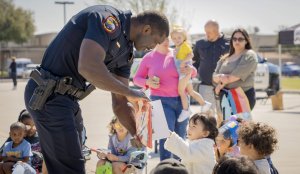 A police officer bends down to hand a book to a young child seated outdoors with other children, while adults and more children look on in the background on a sunny day.
