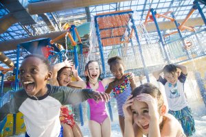 A diverse group of six children is enjoying an indoor water park. They laugh and hold their heads as water splashes down from a play structure behind them. They are wearing colorful swimsuits and are standing under the water sprays with bright smiles.