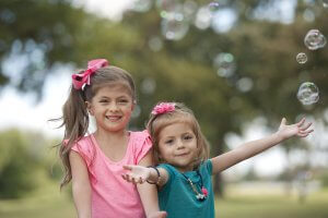 Two young children, both girls, stand in a park. One wears a pink shirt and the other a greenish-blue top. They have big bows in their hair and are smiling at the camera. The girl in green gestures towards floating bubbles around them, with trees blurred in the background.
