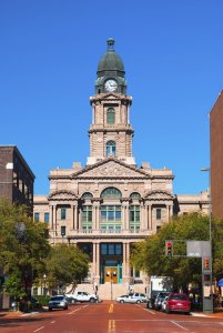 A prominent courthouse stands under a clear blue sky. The building features a clock tower with a green dome, intricate stone architecture, and large arched windows. Vehicles and trees line the brick-paved street leading up to the entrance, flanked by other buildings.
